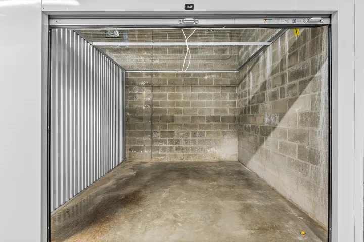 An empty storage unit with cinder block walls, a metal side wall, and a wire mesh ceiling