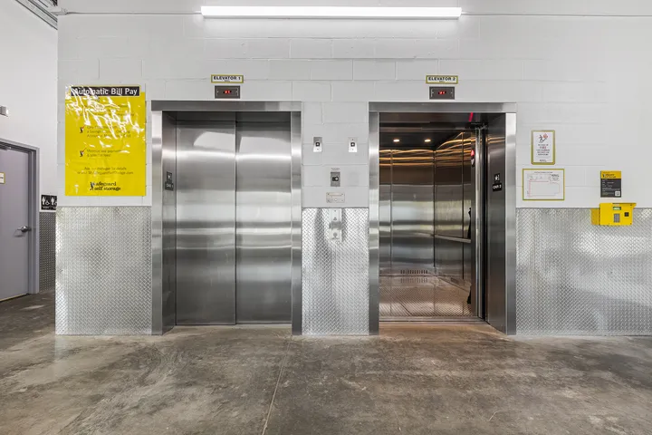 Two stainless steel elevators inside a self-storage facility with a diamond plate wall base and yellow signage
