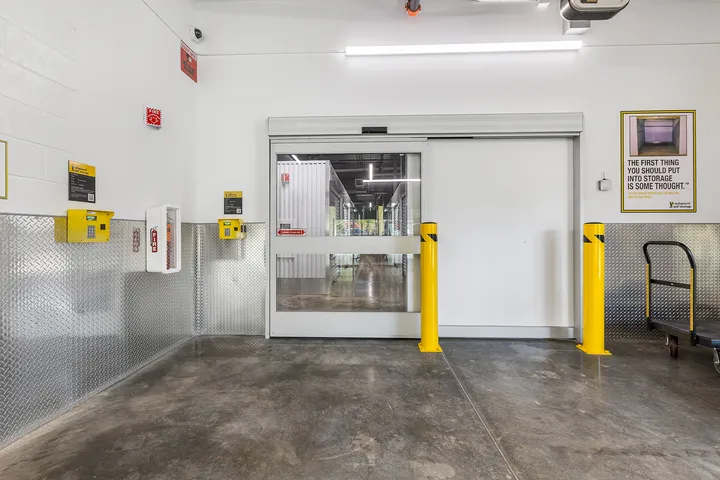 Indoor loading area with a sliding glass door, yellow bollards, keypad, and diamond plate wall panels